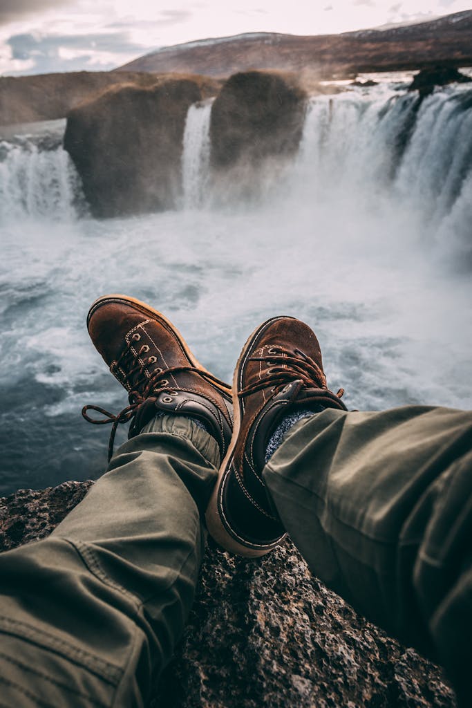 Person in hiking boots sitting at cliff's edge, overlooking breathtaking waterfall.